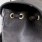 Close-up of a black leather backpack with gold drawstring closures on a light gray background.