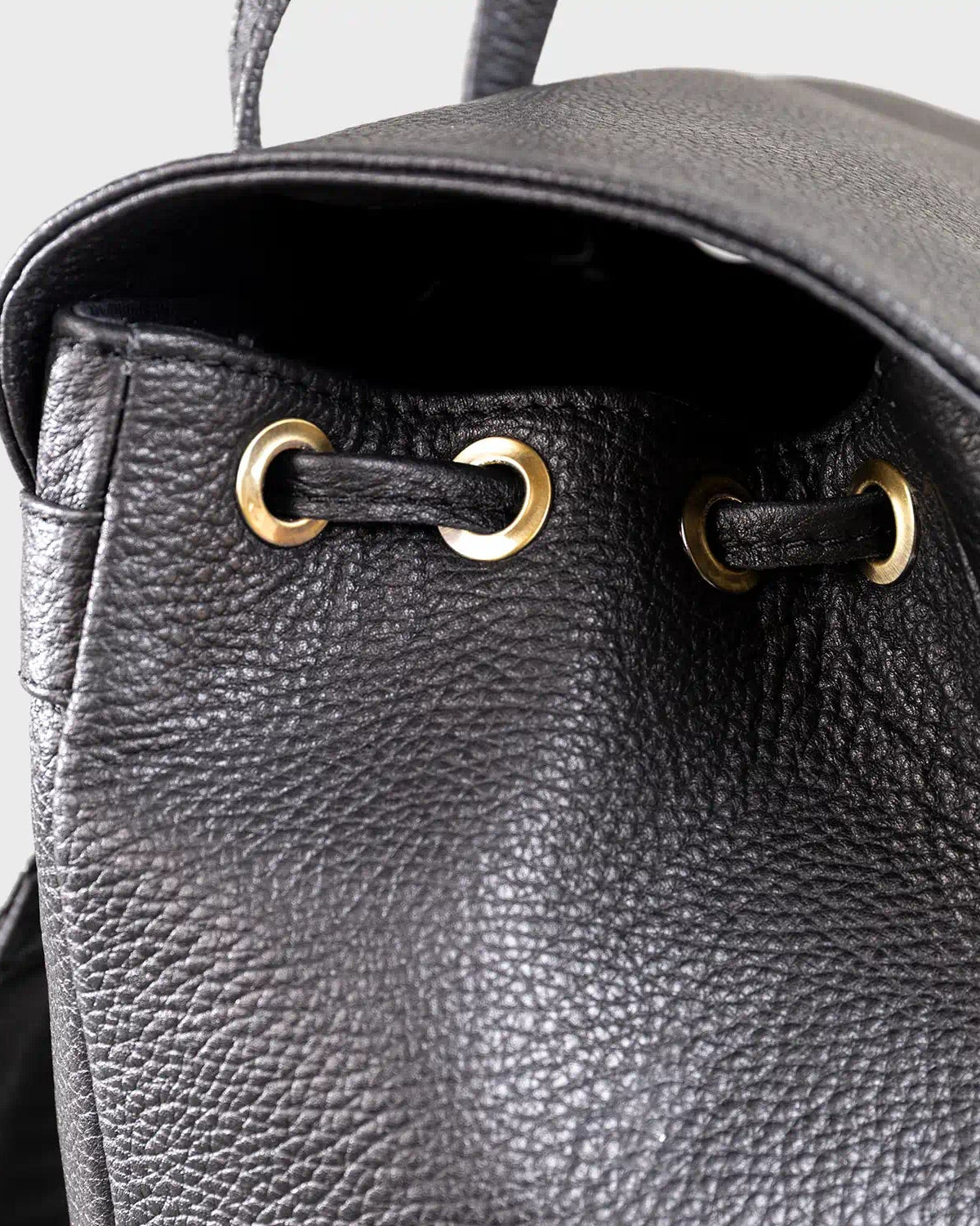 Close-up of a black leather backpack with gold drawstring closures on a light gray background.